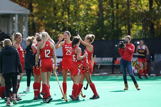 A group of female field hockey players in red uniforms are interacting on a green playing field. One player holds a wooden stick and high-fives a teammate. A woman wearing a red sweatshirt with a camera records the scene. Trees with autumn foliage and a tent are visible in the background.