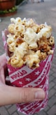 A hand is holding a cone-shaped red and white paper container filled with caramel popcorn. The background features a paved walkway and some greenery, slightly out of focus.