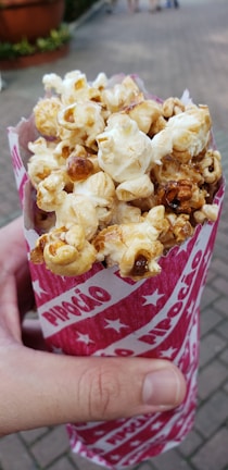 A hand is holding a cone-shaped red and white paper container filled with caramel popcorn. The background features a paved walkway and some greenery, slightly out of focus.