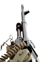 A close-up view of a machine gun from a low angle, focusing on its barrel, and linked ammunition. The gun is metallic with a chain and several bullets attached. The background is plain white, emphasizing the hardware's details.