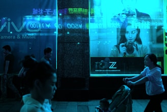 A dimly lit urban scene features people walking at night in front of illuminated store displays. A large advertisement showcases a person holding a camera, set against a vivid blue glow. The store's window reflects the neon signage, adding to the ambiance.
