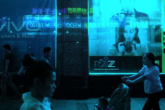 A dimly lit urban scene features people walking at night in front of illuminated store displays. A large advertisement showcases a person holding a camera, set against a vivid blue glow. The store's window reflects the neon signage, adding to the ambiance.