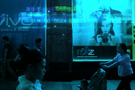 A dimly lit urban scene features people walking at night in front of illuminated store displays. A large advertisement showcases a person holding a camera, set against a vivid blue glow. The store's window reflects the neon signage, adding to the ambiance.