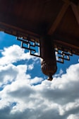 A close-up view of a traditional wooden architectural element is seen against a background of blue sky and fluffy clouds. The intricate carving showcases geometric patterns and details in a dark wood, highlighting craftsmanship.