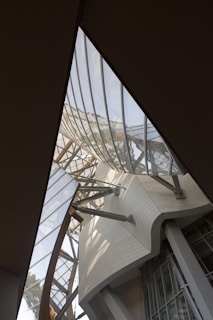 An architectural structure featuring curved and angular glass panels supported by metal beams. The design includes both transparent and opaque materials, with a focus on modern, geometric aesthetics. Sunlight penetrates through the glass, casting intricate shadows.