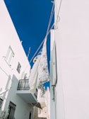 A view of laundry hanging on a line between two white buildings against a clear blue sky. Floral-patterned sheets are gently swaying in the breeze, adding a touch of color and softness against the stark, clean walls. Shadows cast by the buildings create contrasting geometric patterns on the walls.