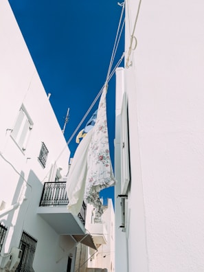 A view of laundry hanging on a line between two white buildings against a clear blue sky. Floral-patterned sheets are gently swaying in the breeze, adding a touch of color and softness against the stark, clean walls. Shadows cast by the buildings create contrasting geometric patterns on the walls.