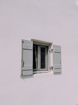 A simple exterior wall painted white with a small window in the center. The window has two light gray shutters partially open, each fitted with metal hinges and a latch. The setting suggests a minimalist architectural style.