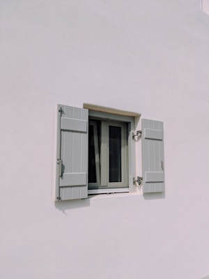 A simple exterior wall painted white with a small window in the center. The window has two light gray shutters partially open, each fitted with metal hinges and a latch. The setting suggests a minimalist architectural style.