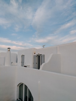 White-washed buildings with blue shutters and doors are set against a backdrop of a clear sky with thin clouds. The architecture is simple yet elegant, typical of Mediterranean style. The smooth, curved edges of the structures blend harmoniously with the sky, creating a serene atmosphere.