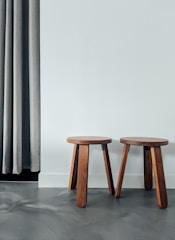 Two wooden stools with simple, round seats and three legs each are placed side by side on a smooth floor. A gray curtain hangs on the left, partially covering the wall.