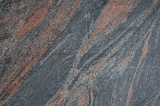 A textured granite surface with swirling patterns of gray and reddish-orange. The composition creates a dynamic and natural appearance with variations in color and texture across the stone.