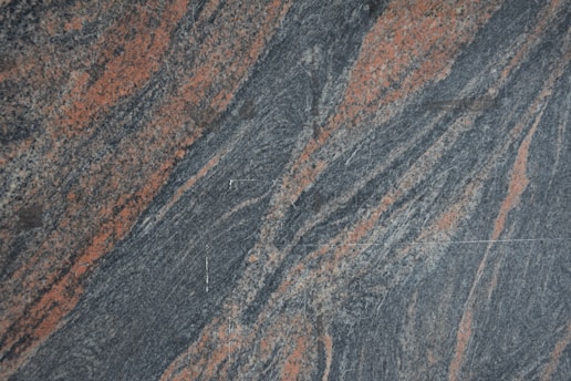 A textured granite surface with swirling patterns of gray and reddish-orange. The composition creates a dynamic and natural appearance with variations in color and texture across the stone.