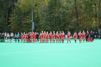 A group of athletes wearing red uniforms stands in a line on a green field hockey pitch. Another team wearing white uniforms stands nearby. The setting is outdoors with a backdrop of lush trees.