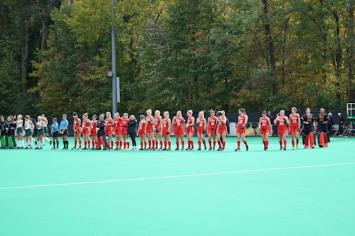 A group of athletes wearing red uniforms stands in a line on a green field hockey pitch. Another team wearing white uniforms stands nearby. The setting is outdoors with a backdrop of lush trees.