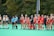 A group of female field hockey players stand on the field, divided into two teams. One team wears white and green uniforms, while the other wears red uniforms. Two referees are present, dressed in blue shirts and black shorts. The players appear to be preparing for a match against a backdrop of lush greenery.