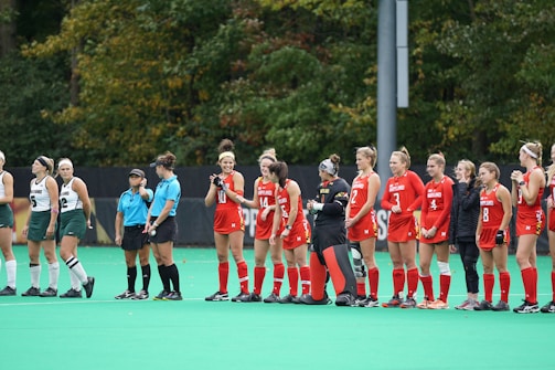 A group of female field hockey players stand on the field, divided into two teams. One team wears white and green uniforms, while the other wears red uniforms. Two referees are present, dressed in blue shirts and black shorts. The players appear to be preparing for a match against a backdrop of lush greenery.