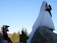 A photographer captures a couple in wedding attire standing on large rocks with a backdrop of trees and a clear blue sky.