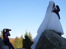 A photographer captures a couple in wedding attire standing on large rocks with a backdrop of trees and a clear blue sky.