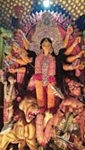 A vibrant and detailed statue depicting a multi-armed goddess adorned with intricate jewelry and colorful garments. She stands triumphantly with a fierce expression, surrounded by a lion and figures representing subjugated demons. The background features elaborate decorations with patterns of intense colors and embellishments.
