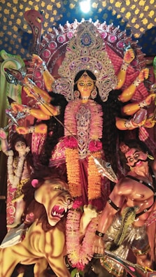 A vibrant and detailed statue depicting a multi-armed goddess adorned with intricate jewelry and colorful garments. She stands triumphantly with a fierce expression, surrounded by a lion and figures representing subjugated demons. The background features elaborate decorations with patterns of intense colors and embellishments.