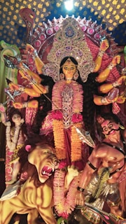 A vibrant and detailed statue depicting a multi-armed goddess adorned with intricate jewelry and colorful garments. She stands triumphantly with a fierce expression, surrounded by a lion and figures representing subjugated demons. The background features elaborate decorations with patterns of intense colors and embellishments.