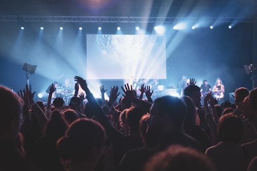 A lively concert scene with a large crowd of people raising their hands in front of a stage. Musicians are performing under bright stage lights, and the atmosphere is energetic and vibrant.