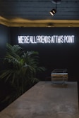 A dimly lit room features a neon sign with the phrase 'WE'RE ALL FRIENDS AT THIS POINT' glowing in white light. Below the sign, there is a marble countertop with a transparent container. To the side, green plants add a touch of nature to the subdued setting.