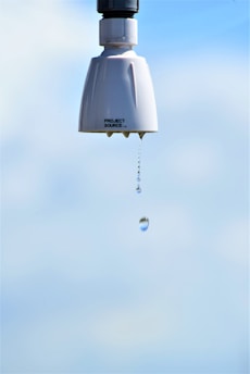 A close-up of a showerhead with water gently dripping from it against a bright blue sky background. The focus is on the water droplet, capturing its descent from the nozzle.