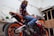 A person wearing a Repsol jacket and jeans sits on a sport motorcycle indoors, surrounded by mirrors and wooden panels. The environment is modern, with neutral tones and some greenery.