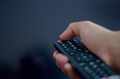 A close-up of a hand holding a remote control against a dark background with red highlights.