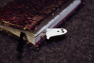 A closed notebook with a sequined cover lies on a textured gray surface. The cover features a mix of red and black sequins. A small, white ghost-shaped bookmark with a smiling face peeks out from the pages.