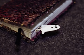 A closed notebook with a sequined cover lies on a textured gray surface. The cover features a mix of red and black sequins. A small, white ghost-shaped bookmark with a smiling face peeks out from the pages.
