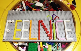 A gray LEGO base plate features a colorful arrangement where LEGO pieces spell out the phrase 'FEEL NICE'. The words are crafted using various colors including white, yellow, orange, red, pink, and green. Behind the plate, there is a collection of assorted loose LEGO bricks in a circular yellow container.