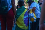 A child draped in a Brazilian flag stands among a group of people. One person is holding a yellow bag with a stuffed bear attached. People are dressed casually, and the scene appears to be outdoors during the day.