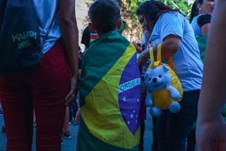 A child draped in a Brazilian flag stands among a group of people. One person is holding a yellow bag with a stuffed bear attached. People are dressed casually, and the scene appears to be outdoors during the day.
