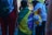 A child draped in a Brazilian flag stands among a group of people. One person is holding a yellow bag with a stuffed bear attached. People are dressed casually, and the scene appears to be outdoors during the day.