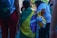 A child draped in a Brazilian flag stands among a group of people. One person is holding a yellow bag with a stuffed bear attached. People are dressed casually, and the scene appears to be outdoors during the day.
