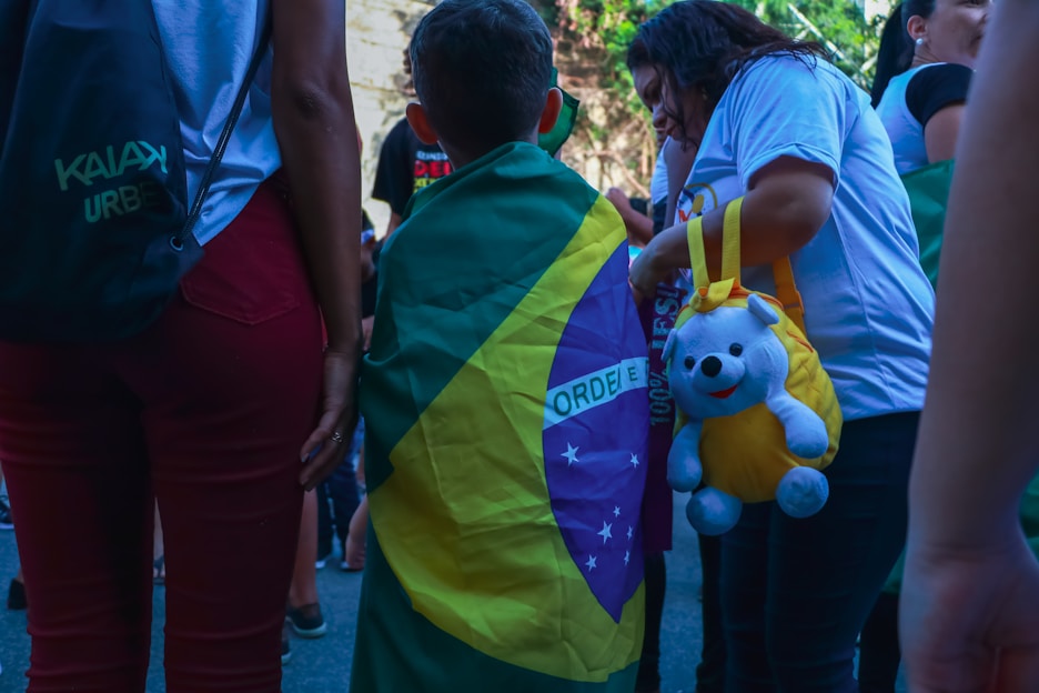 A child draped in a Brazilian flag stands among a group of people. One person is holding a yellow bag with a stuffed bear attached. People are dressed casually, and the scene appears to be outdoors during the day.
