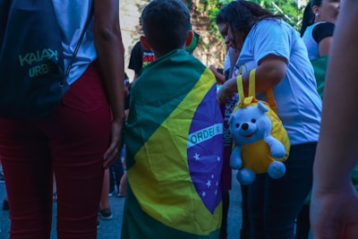 A child draped in a Brazilian flag stands among a group of people. One person is holding a yellow bag with a stuffed bear attached. People are dressed casually, and the scene appears to be outdoors during the day.