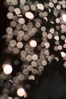An artistic, abstract photograph featuring numerous small light spots, or bokeh, in the foreground set against a dark background. The light spots vary in size and are mostly circular, creating a dreamy and ethereal effect.