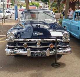 A classic navy blue Chrysler car is parked on a street, with a shiny chrome grille and round headlights. An American flag design is reflected on the side mirror, and a sign is placed in front of the car. Another vintage vehicle is partially visible on the right, and a few people are standing nearby under a shaded area.