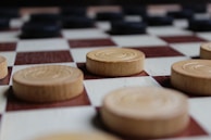 A close-up view of a checkerboard with wooden checkers. The board is arranged in a classic alternating light and dark square pattern. The foreground features light-colored wooden pieces, while darker pieces are visible in the background.