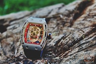 A luxury watch with a skeleton motif on its face is placed on a textured wooden surface. The watch features intricate details, including visible screws and a bold red border surrounding the skull design.