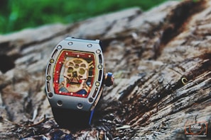 A luxury watch with a skeleton motif on its face is placed on a textured wooden surface. The watch features intricate details, including visible screws and a bold red border surrounding the skull design.