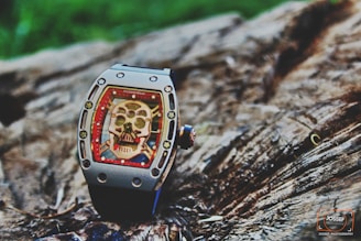 A luxury watch with a skeleton motif on its face is placed on a textured wooden surface. The watch features intricate details, including visible screws and a bold red border surrounding the skull design.