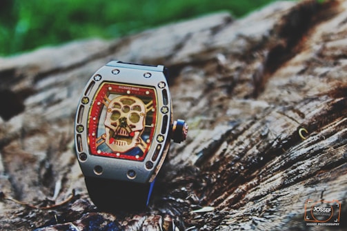 A luxury watch with a skeleton motif on its face is placed on a textured wooden surface. The watch features intricate details, including visible screws and a bold red border surrounding the skull design.