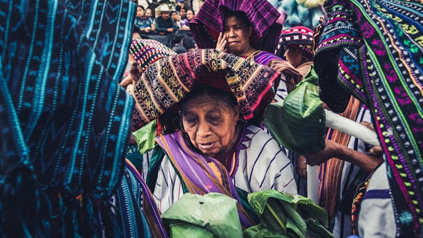 A group of people are dressed in vibrant, traditional attire adorned with intricate patterns. They appear to be participating in a cultural or ceremonial event. The foreground shows an elderly figure wearing a headdress, holding green leaves, surrounded by others also wearing traditional clothes.