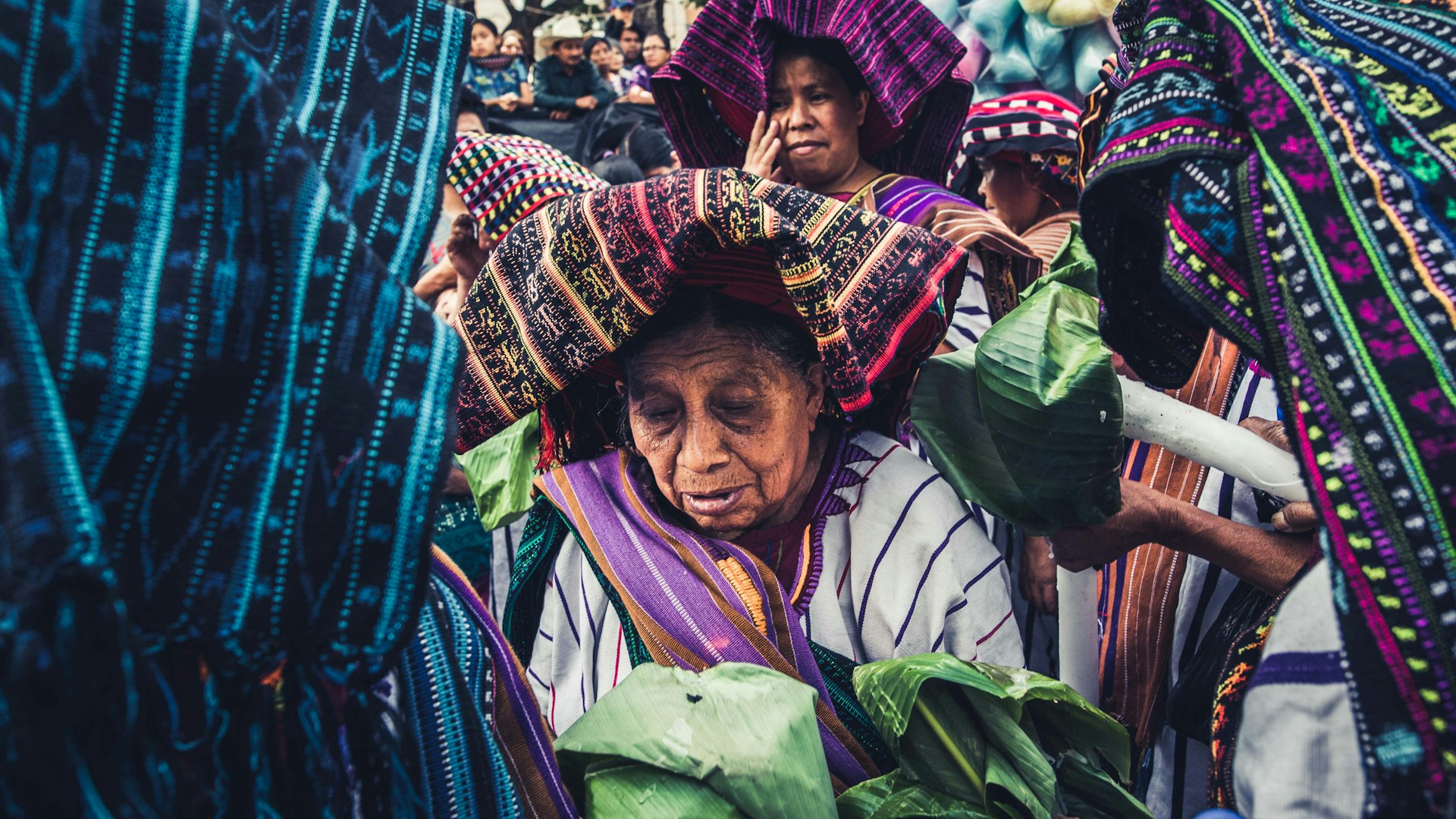 A group of people are dressed in vibrant, traditional attire adorned with intricate patterns. They appear to be participating in a cultural or ceremonial event. The foreground shows an elderly figure wearing a headdress, holding green leaves, surrounded by others also wearing traditional clothes.