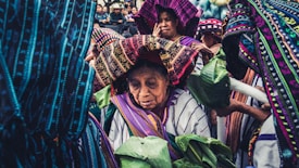 A group of people are dressed in vibrant, traditional attire adorned with intricate patterns. They appear to be participating in a cultural or ceremonial event. The foreground shows an elderly figure wearing a headdress, holding green leaves, surrounded by others also wearing traditional clothes.